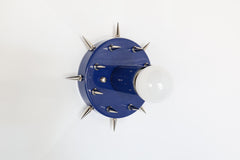 Side-angle product photo of the Sid Sconce in glossy navy with silver-tone spikes, mounted as a small wall sconce against a simple white background. The image shows the fixture’s dimensional shape, projecting bulb, spiked round backplate, and bold mix of playful color with sharper metal accents.