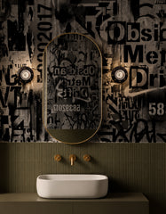 A pair of black and gold Sid Sconces installed on either side of a brass-framed oval mirror in a dramatic powder room. The space features black-and-white graphic wallpaper, olive green ribbed wall tile, a modern white vessel sink, and warm brass plumbing fixtures for a bold, moody, punk-inspired look.