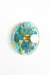 Modern two light wall sconce with brass hardware and blue flowers. Features resin-encapsulated orange flowers with greenery, ferns, and leaves.  Adds a real-like nature theme to any interior design project.  Called the Botanico Sconce by Sazerac Stitches. Features Bkue flowers with greenery