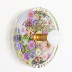 Modern two light wall sconce with brass hardware and purple flowers. Features resin-encapsulated green flowers with greenery, ferns, and leaves.  Adds a real-like nature theme to any interior design project.  Called the Botanico Sconce by Sazerac Stitches. Features green flowers with greenery