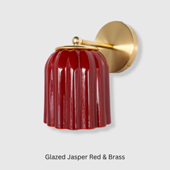 The Marcella Sconce pairs a richly glazed, fluted shade in jasper red with a simple brass arm and backplate, giving it real depth of color without needing a busy silhouette to stand out. The ribbed, scalloped-edge shade catches light differently from every angle, making it a strong choice for a bathroom vanity, powder room, or entryway that wants a warm, tactile fixture instead of a plain glass shade.