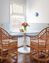 Bright kitchen breakfast nook with built-in striped banquette seating, round white pedestal table, and woven rattan dining chairs on warm wood floors, styled with a vase of pink tulips and a patterned Roman shade at the window. On the wall is the Sky Blue ceramic Lierre Sconce by Sazerac Stitches, a glossy botanical leaf-style ceramic wall light with a frosted globe, adding grandmillennial coastal charm and soft designer lighting.