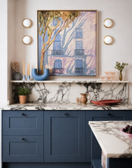 Artful slate blue kitchen with two stacked pairs of Abeille sconces framing a large painterly wall piece above a marble shelf, marble backsplash, and matching marble countertop. The round white globe lights and warm metal trim add soft symmetry against the pale wall, while the blue cabinetry, black knobs, blush glassware, sculptural vessels, and framed artwork create a polished modern traditional kitchen with a collected, gallery-like feel. A strong fit for kitchen wall sconces, blue kitchen cabinets, marbl
