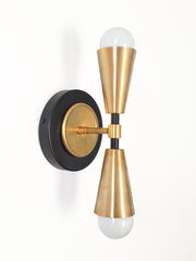 A small black and brass wall sconce with two raw brass cones that meet at a black collar, shaped like an hourglass. A brass disc sits inside a black backplate ring, so the layers read clean and graphic. It lights up and down, giving a soft wash above and below. Use it as a vanity light beside a bathroom mirror or a pair in a powder room. Black and brass look sharp on white walls and patterned tile, a good fit for mid century modern and art deco bathroom lighting.