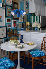 A small kitchen corner packed with personality: turquoise walls disappear behind a gallery wall of art and ephemera, bamboo chairs in blue batik fabric surround a white tulip table, and a cobalt blue fluted pendant light on brass hangs low over the setting. Green and white stripes, a yellow stand mixer, and a blue and white vase of hydrangeas round out the color story. It’s collected eclectic at its most joyful, proof a tiny kitchen can carry a huge amount of color.