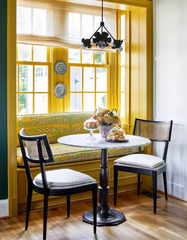 Bright breakfast nook featuring the Small Octavia chandelier in all black, hanging above a marble bistro table with a pedestal base and black dining chairs with cane backs. The cozy dining corner includes bold yellow window trim, patterned banquette seating, hardwood floors, and vintage decorative plates, blending eclectic cottage charm with modern statement lighting.