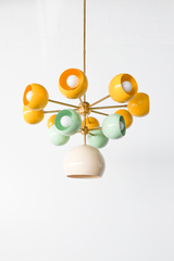 Angled view of the Solaris chandelier, a modern sputnik chandelier with branching brass arms and glossy orb-shaped shades in sunshine yellow and soft mint green, plus a cream central dome shade, designed for design-forward homeowners who love bold color, mid century modern interiors, and artful statement lighting over a dining table, kitchen island, or entryway.