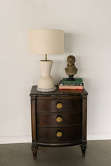 Dark Nightstand with a modern cream lamp by Sazerac Stitches and brass sun-shaped cast brass drawer pulls.  Antique books and a vintage bust of Napoleon.