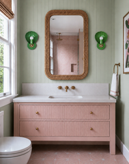 A pink and sage green bathroom that feels fresh and a little old-fashioned in the best way: V-groove paneled walls in soft sage, a blush vanity with fluted drawer fronts and brass knobs, a pink star-pattern tile floor and a woven rattan mirror. A pair of green shell-shaped sconces with bare white globe bulbs flank the mirror, adding a punch of saturated color. Collected, feminine and fully committed to color.