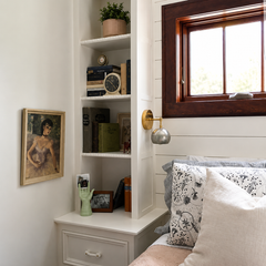 Small bedroom nook featuring a compact Loa wall sconce with a steel globe shade and warm brass backplate, mounted beside a wood-trimmed window above the bed. The minimal mixed-metal sconce adds a soft bedside glow and a sculptural accent, pairing beautifully with white shiplap walls, built-in shelves, and patterned pillows. Ideal for bedside wall sconce lighting, reading nook lighting, and modern farmhouse or cottage bedroom decor.