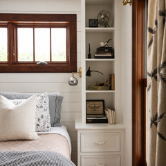Bright bedroom vignette with a compact Loa wall sconce in mixed metals, featuring a brushed steel globe shade and warm brass backplate, mounted on a white horizontal shiplap wall between a wood-trimmed window and built-in shelving. The small modern globe sconce provides focused bedside reading light without taking up nightstand space, pairing with crisp white built-ins, vintage-inspired decor, and neutral bedding with black-and-white striped textiles. Great for modern farmhouse bedroom lighting, cottage bed