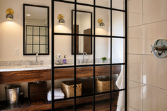 Modern bathroom vanity scene with a pair of Loa wall sconces featuring brushed brass round backplates and steel dome shades, casting warm downward light beside two black framed mirrors. Styled with a floating wood vanity, marble countertop, woven storage baskets, and a black windowpane grid glass shower partition for an industrial modern look. Great inspiration for bathroom vanity lighting, bedside sconces, modern brass and steel wall lights, minimalist lighting, and warm accent lighting in contemporary int