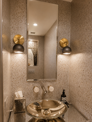 Warm, modern powder room with a tall rectangular mirror flanked by two Loa wall sconces in smoky steel and brass. The compact globe shades cast a soft downlight onto textured neutral stone walls, creating a boutique hotel vibe. A sleek small bathroom sconce idea for limestone, travertine, microcement, or plaster bathrooms with warm brass fixtures and minimalist styling.
