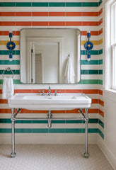 Lagoon and brass Medium Thalia Sconces shown as a matching pair of vertical bathroom vanity lights on each side of a large rounded rectangular mirror. The colorful bathroom features glossy striped wall tile in teal, green, coral, mustard, and cream, a vintage-style white console sink with chrome legs, white hex floor tile, and bright natural light. A playful two-light wall sconce for kids bathrooms, family bathrooms, colorful powder rooms, preppy bathrooms, striped tile bathrooms, and cheerful retro-inspire