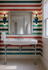 Lit Lagoon and brass Medium Thalia Sconces installed beside a bathroom mirror in a bold striped tile bathroom with green, teal, coral, yellow, and cream bands. The vertical double-bulb sconces add warm light while keeping the room playful, polished, and functional. Great inspiration for colorful bathroom lighting, kids bathroom sconces, guest bath vanity lights, retro bathroom design, preppy interiors, and bright powder room lighting.