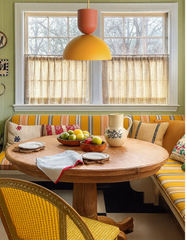 Taeuber Pendant Light in mustard yellow and terra cotta with brass stem, styled over a vintage-inspired breakfast nook with striped yellow banquette seating, oak round pedestal dining table, cane chair, and patterned throw pillows. Warm and colorful kitchen nook with mid-century modern and retro influences, accented by green paneled walls, patterned café curtains, and eclectic cottage details. Handmade two-tone pendant lighting by Sazerac Stitches — perfect for kitchens, dining rooms, and bold breakfast noo