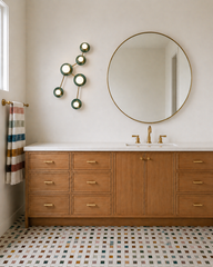 Neutral bathroom with warm oak vanity and white countertop, a large round brass-framed mirror, and brass faucet hardware and towel bar. A multicolor mosaic floor tile mixes cream, sage, mustard, and muted tones for a custom designer finish. Mounted beside the mirror is the Sazerac Stitches Taurus sconce, a constellation-inspired wall sculpture light in Mermaid Green discs with brass arms and globe bulbs, styled as modern vanity lighting and artful wall decor. 