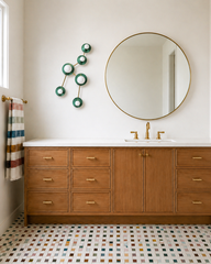 Neutral bathroom with warm oak vanity and white countertop, a large round brass-framed mirror, and brass faucet hardware and towel bar. A multicolor mosaic floor tile mixes cream, sage, mustard, and muted tones for a custom designer finish. Mounted beside the mirror is the Sazerac Stitches Taurus sconce, a constellation-inspired wall sculpture light in Mermaid Green discs with brass arms and globe bulbs, styled as modern vanity lighting and artful wall decor. Keywords: green bathroom lighting, brass and gre