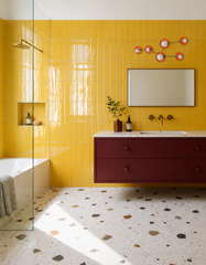 A bold, design-forward bathroom with glossy sunshine yellow wall tile installed in a vertical stacked pattern, creating a bright color-drenched shower and vanity backdrop with tons of reflective texture. A floating burgundy vanity adds rich contrast, paired with a minimalist rectangular mirror, warm brass faucet and shower fixtures, and a terrazzo-style floor with multicolor stone flecks for a modern, high-end finish. Mounted above the mirror is the Sazerac Stitches Taurus sconce, a constellation-inspired s