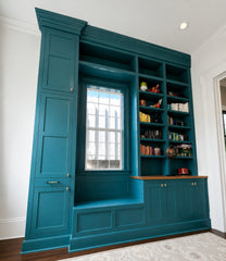 Color-drenched teal built-in cabinetry creates a custom reading nook with a window seat, paneled storage doors, open bookshelves, brass hardware, and a warm wood counter. The room feels tailored and traditional but still bold, with deep blue-green millwork turning a plain wall into a library-style storage moment. This image works for searches like teal built-ins, custom bookshelves, window seat ideas, home library nook, traditional millwork, colorful cabinetry, reading nook storage, and maximalist family ro