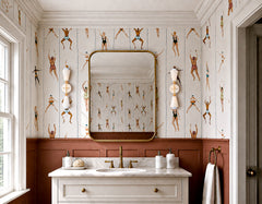 Playful traditional bathroom with cream and brass Janus sconces flanking a large brass-framed mirror over a marble vanity. Vintage swimmer wallpaper, terra cotta paneled wainscoting, brass faucet, cream cabinetry, and classic molding give the room a polished but quirky grandmillennial feel. The geometric two-light sconces add sculptural personality and work beautifully for a kids bathroom, powder room, or maximalist bath with vintage sports wallpaper and warm brass vanity lighting.