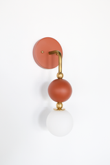 The Ida Sconce in terra cotta stacks a hooked brass arm, small brass ball joints, and a matte terra cotta accent ball above a soft white opal glass globe, giving the whole fixture a warm, sculptural silhouette on the wall. It works well as a bedside light in place of a table lamp, beside a bathroom mirror, or anywhere a single statement sconce makes more sense than a matching pair.
