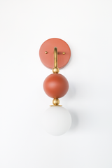 There's a lot going on in a small footprint with the Ida Sconce: a hooked brass arm drops down through two small brass balls to hold a matte terra cotta sphere, then continues to a soft, frosted glass globe at the bottom. The earthy terra cotta tone and warm brass hardware make it feel more like a piece of jewelry than standard bathroom lighting, at home beside a bed, above a vanity, or anywhere that could use one confident, sculptural fixture instead of a plain sconce.
