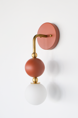 There's a lot going on in a small footprint with the Ida Sconce: a hooked brass arm drops down through two small brass balls to hold a matte terra cotta sphere, then continues to a soft, frosted glass globe at the bottom. The earthy terra cotta tone and warm brass hardware make it feel more like a piece of jewelry than standard bathroom lighting, at home beside a bed, above a vanity, or anywhere that could use one confident, sculptural fixture instead of a plain sconce.
