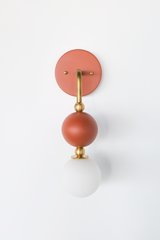 The Ida Sconce in terra cotta stacks a hooked brass arm, small brass ball joints, and a matte terra cotta accent ball above a soft white opal glass globe, giving the whole fixture a warm, sculptural silhouette on the wall. It works well as a bedside light in place of a table lamp, beside a bathroom mirror, or anywhere a single statement sconce makes more sense than a matching pair.