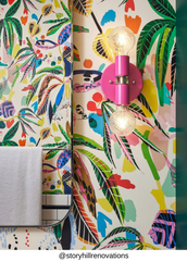 Close view of small Thalia wall sconce in doll pink with brass hardware and two glowing globe bulbs, installed on vibrant tropical patterned wallpaper, a bold maximalist bathroom lighting choice for powder rooms, colorful interiors, and statement wall sconce styling.