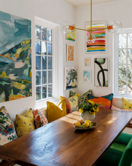A colorful maximalist dining room layers abstract artwork, patterned pillows, a built-in window banquette, green velvet chairs, and a long rustic wood table styled with sunflowers and citrus. Above it, the customizable Three-Light Cadiz Chandelier by Sazerac Stitches pairs clear glass globes with Orange sockets and Brass hardware for playful modern statement lighting. The airy fixture complements the room’s gallery wall without competing with its saturated colors and mixed patterns. Explore colorful dining 