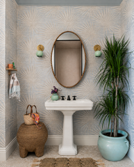 Beachy powder room featuring mint green Loa wall sconces in brass flanking a rattan-framed oval mirror over a white pedestal sink. Tropical palm fan wallpaper, woven stool, textured rug, and a large indoor plant add coastal boho style. Strong SEO for bathroom vanity sconces, coastal bathroom lighting, mint brass sconce, and tropical powder room decor.
