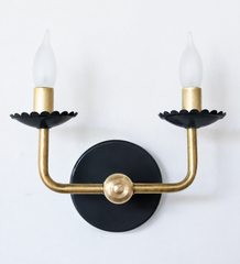 A classic two-arm candle sconce goes moodier here, with scalloped bobeche cups in matte black beneath frosted flame-tip bulbs and warm brass arms. It keeps the traditional shape but leans more serious and graphic, a good fit for a library, dining room, or any space that wants classic lighting without the sweetness.