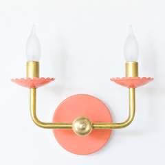 A simple, modern U-shaped arm in raw brass carries a softer color story here, with scalloped bobeche cups and a backplate in matte peach beneath frosted flame-tip bulbs. It keeps the traditional candle-sconce detail but trades a bold primary for something gentler and sun-faded, the kind of sconce that would sit easily in a bathroom, hallway, or bedroom that wants color without shouting.