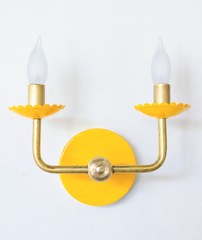 A classic two-arm candle sconce turns cheerful here, with scalloped bobeche cups in a saturated yellow beneath frosted flame-tip bulbs and traditional curved brass arms. It brings sunny, colorful energy to a familiar shape, well suited to a breakfast nook or kitchen that wants personality.