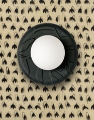 Close-up of the Unglazed Black Lierre ceramic sconce by Sazerac Stitches, featuring a matte black porcelain wall plate with sculptural raised leaf-and-vine relief and a smooth white globe diffuser. Styled against creamy neutral and black mini flower wallpaper with a repeating black floral bud motif, this modern botanical light adds high-contrast drama and artisanal texture for moody powder rooms, grandmillennial spaces, and chic French-inspired interiors. Adds an artisanal, handmade touch.