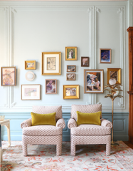 French-inspired living room with pale blue paneled walls and ornate plaster molding, styled with an eclectic gallery wall of vintage art in gold frames above two patterned club chairs with chartreuse velvet lumbar pillows. A soft floral area rug and a brass palm floor lamp with a frosted globe add warm, traditional European charm. On the wall, the Unglazed Porcelain Lierre Sconce by Sazerac Stitches adds sculptural botanical texture and soft ambient lighting, a refined ceramic wall light for grandmillennial