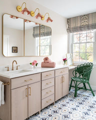 Light and bright bathroom with light wood cabinetry, three brass and peach sconces, brass framed mirror, and patterned penny tile floor