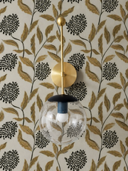 A brass and navy wall sconce brings a little whimsy to this botanical moment, its clear glass globe catching light against gold and black leaf wallpaper with dotted flower clusters. The ball finial on top and the dark cap over the glass give it a collected, slightly playful shape, the kind of colorful, grandmillennial-leaning lighting that pairs well with vintage furniture, patterned wallpaper, and rooms that already have a point of view.