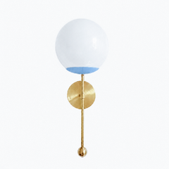 A fresh sky-blue globe sconce for a powder room, bedside, or hallway that wants a wall light with a little Southern charm. The Verbena Sconce tops a round brass backplate with a slim brass stem, seats a white glass globe in a soft celestial-blue cup, and finishes the bottom with a small brass ball, the kind of detail that makes it feel custom. Light blue and brass together read airy and classic, at home flanking a mirror, art, or a bed, especially in a coastal or Southern traditional space. A soft, glowing 