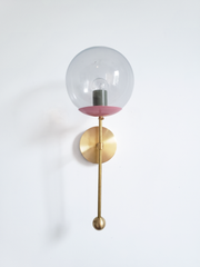A color-mixed globe sconce for a powder room, bedside, or hallway that wants a wall light with a genuinely custom feel. This Verbena Sconce swaps the usual opaque shade for a clear glass globe, so the bulb shows through, seated in a blush-pink cup with an olive socket sleeve inside for an unexpected two-tone pairing. A slim brass stem, round backplate, and small brass ball finish it below. The pink-and-olive combination reads playful and collected, the kind of offbeat choice that makes people ask where you 