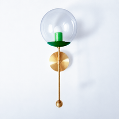 A tonal green globe sconce for a powder room, bedside, or hallway that wants a wall light with a genuinely custom feel. This Verbena Sconce swaps the usual opaque shade for a clear glass globe, so the bulb shows through, seated in a deep mermaid-green cup with a brighter spring-green socket sleeve inside for a considered green-on-green pairing. A slim brass stem, round backplate, and small brass ball finish it below. The layered greens read fresh and collected, the kind of thoughtful detail that makes peopl