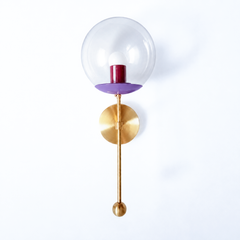 A rich jewel-tone clear-globe sconce for a powder room, bedside, or hallway that wants a wall light with a genuinely custom feel. This Verbena Sconce swaps the usual opaque shade for a clear glass globe, so the bulb shows through, seated in an orchid-purple cup with a deep black-cherry socket sleeve inside for a saturated two-tone pairing. A slim brass stem, round backplate, and small brass ball finish it below. The purple-and-berry combination reads bold and a little sultry, the kind of considered detail t