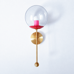 A high-energy color-mixed globe sconce for a powder room, bedside, or hallway that wants a wall light with a genuinely custom feel. This Verbena Sconce swaps the usual opaque shade for a clear glass globe, so the bulb shows through, seated in a bright red cup with a hot-pink socket sleeve inside for a fearless red-and-pink pairing. A slim brass stem, round backplate, and small brass ball finish it below. The red-and-pink combination reads playful and bold, the kind of unexpected choice that makes people ask