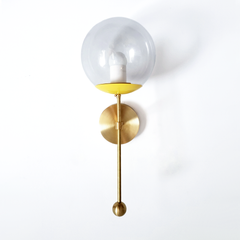 A cheerful clear-globe sconce for a powder room, kitchen, or bedside that wants a wall light with a custom feel and a hit of sunshine. This Verbena Sconce swaps the usual opaque shade for a clear glass globe, so the bulb shows through, seated in a sunny yellow cup with a clean white socket sleeve inside. A slim brass stem, round backplate, and small brass ball finish it below. The yellow-and-white pairing reads bright and easy, warmer than a single accent and lovely flanking a mirror, art, or a bed. A glowi