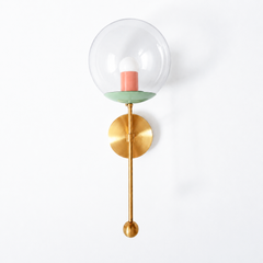 A soft sherbet-toned clear-globe sconce for a powder room, bedside, or hallway that wants a wall light with a genuinely custom feel. This Verbena Sconce swaps the usual opaque shade for a clear glass globe, so the bulb shows through, seated in a fresh vista-green cup with a warm peach socket sleeve inside for a gentle, unexpected pairing. A slim brass stem, round backplate, and small brass ball finish it below. The green-and-peach combination reads fresh and a little nostalgic, the kind of considered detail