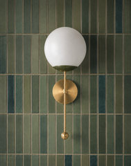 A glossy opal glass globe sits above a brass backplate on this wall sconce, with a slim rod and ball pull finishing the line below. Set against narrow vertical tile in shifting sage and teal tones, it has a calm, spa-like feel that suits a zen bathroom, shower wall, or vanity area where the tile is doing as much of the design work as the fixture.