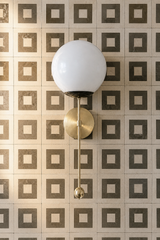 A round opal glass globe glows above a brass backplate on this wall sconce, with a slim brass rod and ball pull finishing the look below. Set against warm square tile in a bronze grid pattern, it reads polished with a little Art Deco edge, the kind of brass lighting that suits a bathroom vanity wall or powder room with graphic tile on the floor or walls.