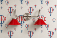 Two-light vanity light with red gloss cone shades on curved nickel gooseneck arms and a round nickel backplate. Put it over a bathroom mirror or use it as a double wall sconce over a desk or bunk beds. Here it sits on off-white wallpaper printed with red and blue hot air balloons, so the red shades pick up the print. It suits kids bathroom lighting, a playful nursery or playroom, and red white and blue rooms with a vintage, slightly nautical feel.