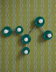 Graphic olive-green wallpaper with a repeating pale blue chain motif creates a bold patterned setting for the Mermaid Green Virgo wall sconce with Chrome hardware. Glossy teal-green concave shades and white globe bulbs follow the Virgo zodiac constellation, giving the six-light fixture the presence of functional wall art. This tonal green interior combines retro-inspired pattern, celestial design, sculptural lighting, and color-drenched maximalism. Find customizable statement lighting by Sazerac Stitches.